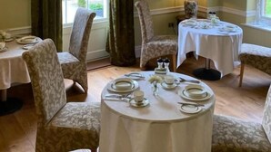 A dining area with two round tables set for four, each with white tablecloths and silverware. There are two upholstered chairs with a floral pattern. A chandelier hangs from the ceiling, and a window with green curtains allows natural light into the room. - Reymerston Hall (Norwich)