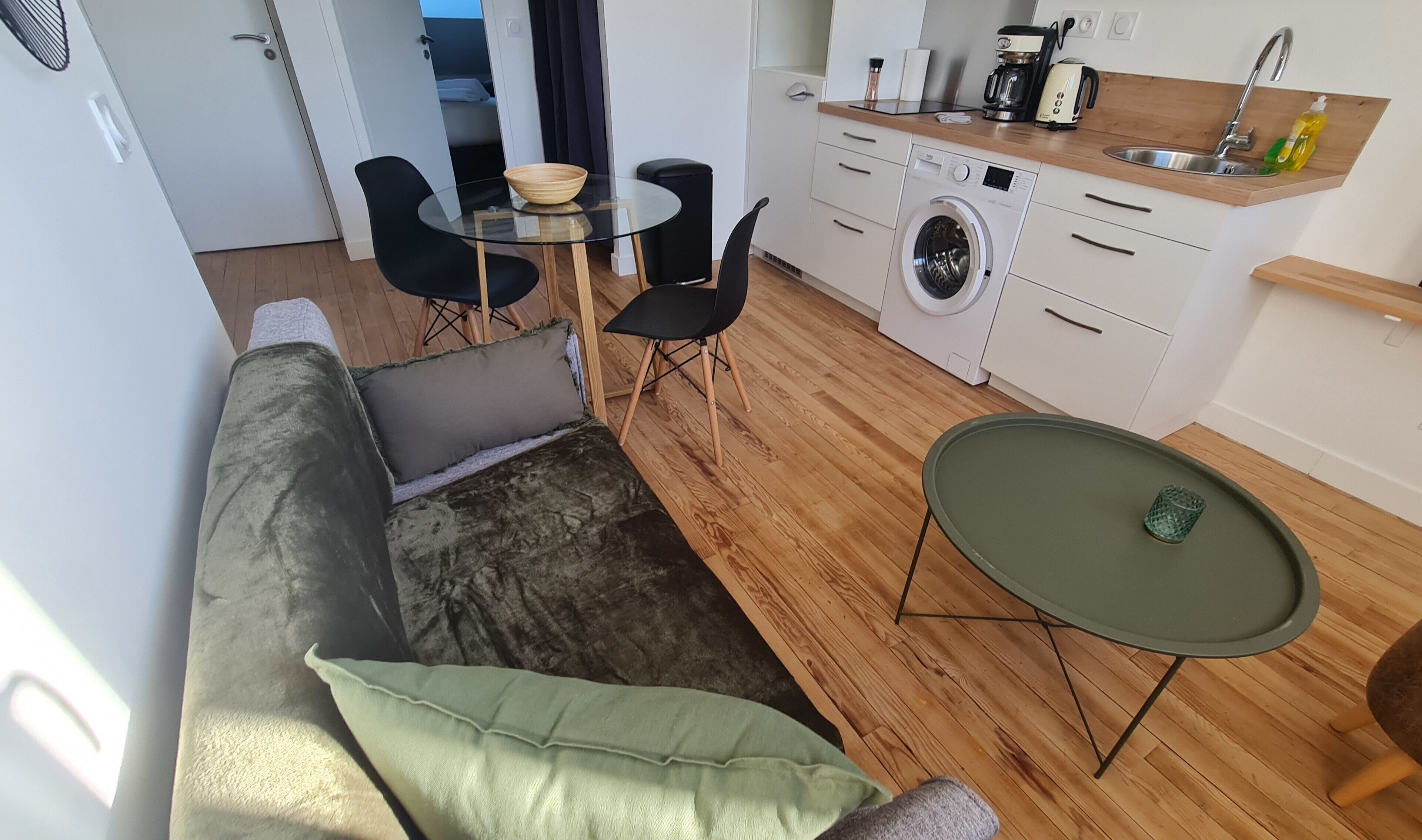 A compact living space with a sofa, a small round table, a washing machine, and a kitchenette.