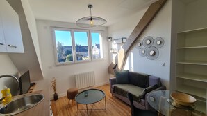 Ett kompakt bostadsutrymme med en soffa, ett litet bord, ett element, ett fönster med utsikt och en köksdel med en diskho och ett diskställ.