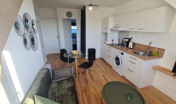 A compact kitchen with a washing machine, a coffee maker, and a sink. There is a dining area with a table and chairs.