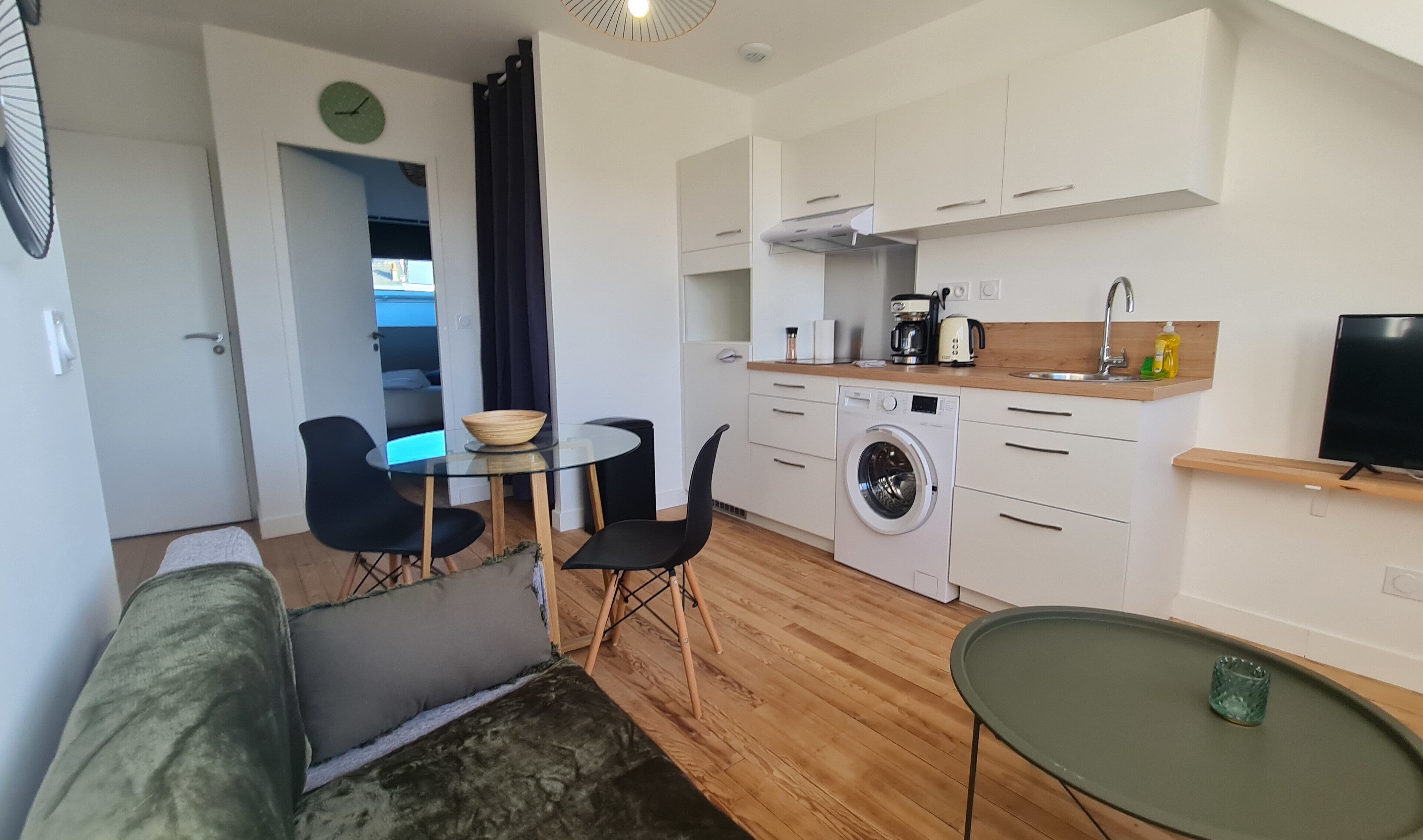 A compact kitchen with a washing machine, a dining area with a glass table, and a living area with a sofa.