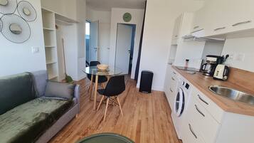 A compact living space with a sofa, a dining table with chairs, a washing machine, and a kitchenette with a coffee maker and toaster.