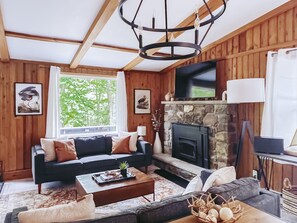 A cozy living room with a fireplace, wooden walls, and a large window. - Chic Poconos Home: Fall Foliage Escape (Clifton Township)