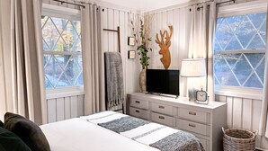 Ett sovrum med en säng, en tv, en byrå, en lampa och ett fönster med utsikt över träd.