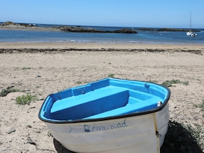 Una pequeña embarcación con interior azul, identificada con la leyenda "Portread", en una playa arenosa con mar tranquilo y rocas al fondo.