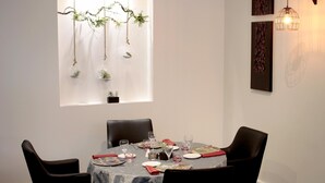 Un coin repas avec une table ronde dressée pour quatre personnes, entourée de chaises noires. La table est recouverte d’une nappe à motifs, et on y trouve des assiettes, des verres et des couverts.