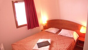 A single bed with a striped bedspread, two bedside lamps, a window with red curtains, and a red headboard.