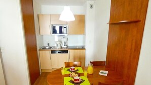 A compact kitchen with a microwave, coffee maker, and sink. A dining table with chairs is set for two, with plates, cups, and a glass of orange juice.