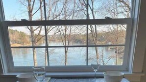 Una mesa de comedor con dos sillas, una ventana con vistas a árboles y agua, y un mantel individual con un cuenco y una copa de vino.