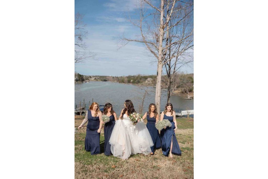 A group of women in formal attire standing by a body of water with a tree and a fence in the background.
