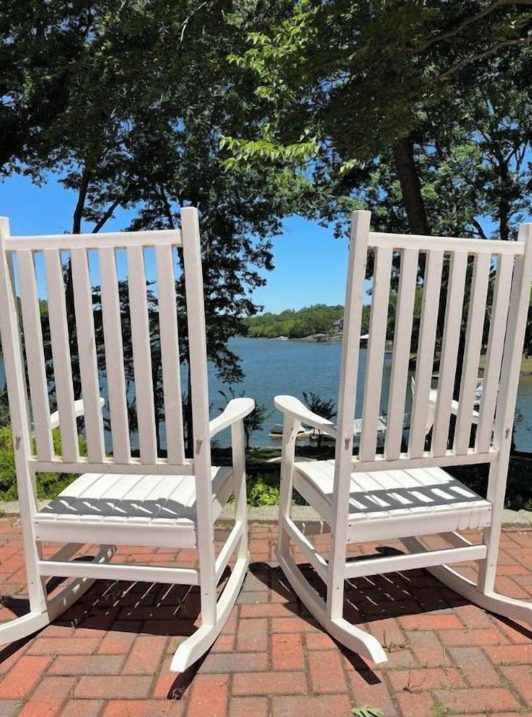 Two white Adirondack chairs facing a body of water with trees in the background.
