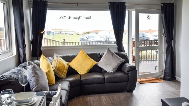 A modern living room with a black leather sofa, a dining table with plates and a wine glass, and a view of a parking area through a glass door.