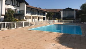 Ein Schwimmbecken, umgeben von einem weißen Zaun, mit einem Gebäude im Hintergrund, das Balkone und ein Ziegeldach hat.