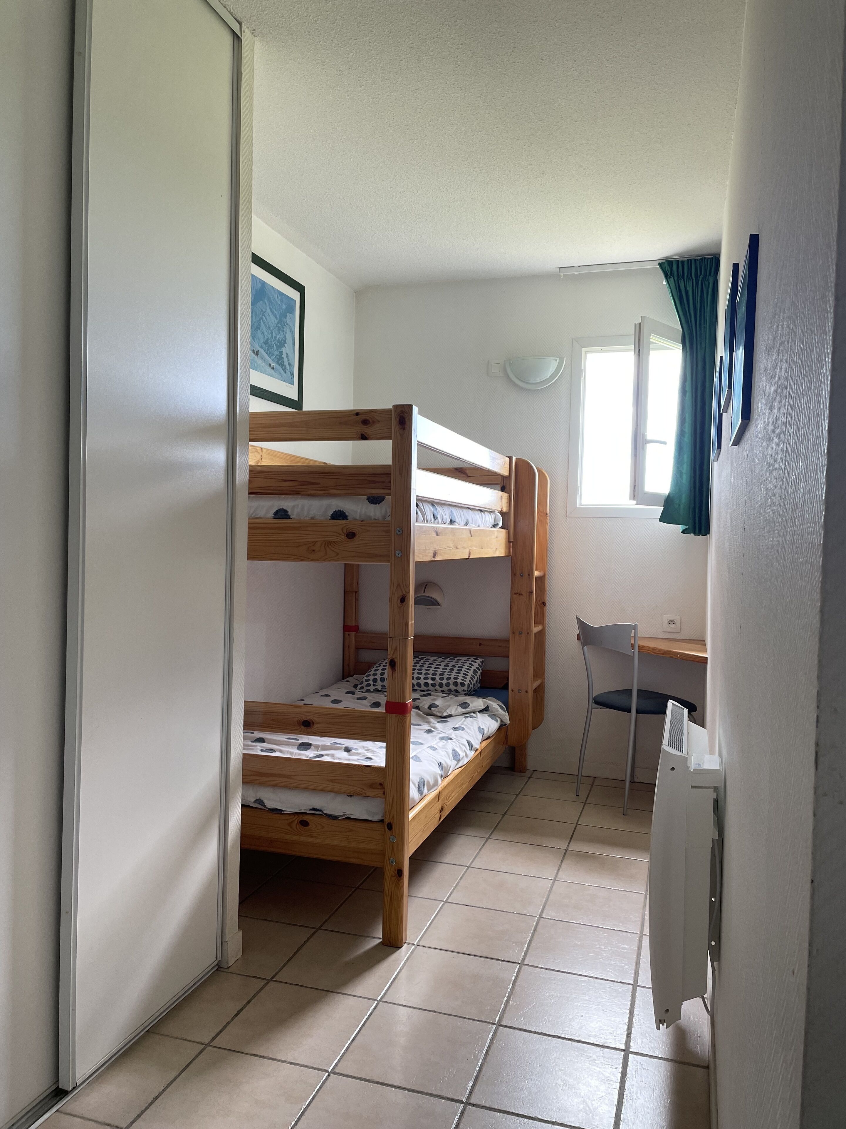 A dormitory room with bunk beds, a desk, a chair, and a radiator.
