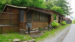 A wooden cabin with a porch, a small front yard with rocks and plants, and a gravel path leading to the road. - Riverside Suite*HotTub*WiFi*Steps to the River (Elkins)