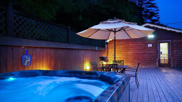 A wooden deck with a hot tub, an umbrella, and a small building with a door. - Riverside Suite*HotTub*WiFi*Steps to the River (Elkins)