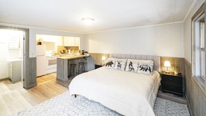 A bedroom with a large bed, a nightstand with a lamp, and a view of the kitchen. - Riverside Suite*HotTub*WiFi*Steps to the River (Elkins)
