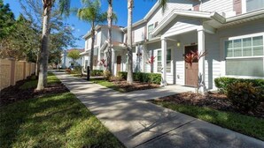 A row of white two-story houses with a covered front porch, palm trees, and a clear blue sky.