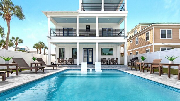 A pool area with lounge chairs, a dining table, and a two-story building with balconies. - Nautilus! Sleeps 22! Ocean View! Rooftop Deck (Destin)