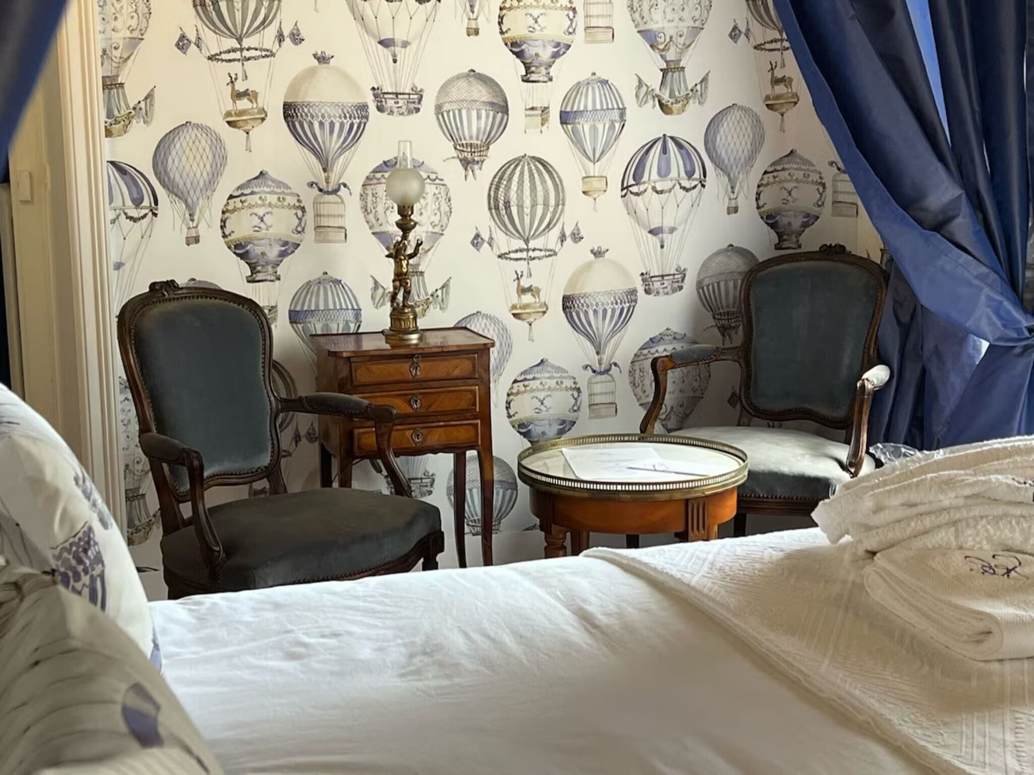 A room with a patterned wallpaper featuring hot air balloons, a wooden side table with a lamp, two upholstered chairs, and a bed with white linens.