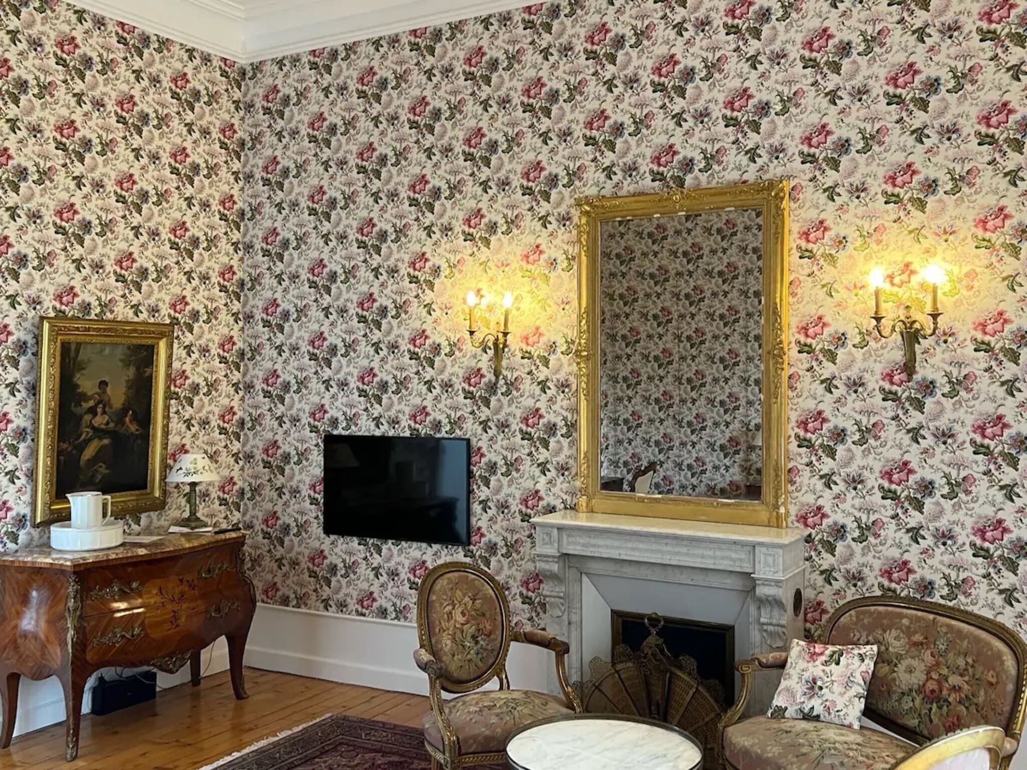 A room with floral wallpaper, a fireplace, a television, and antique furniture.