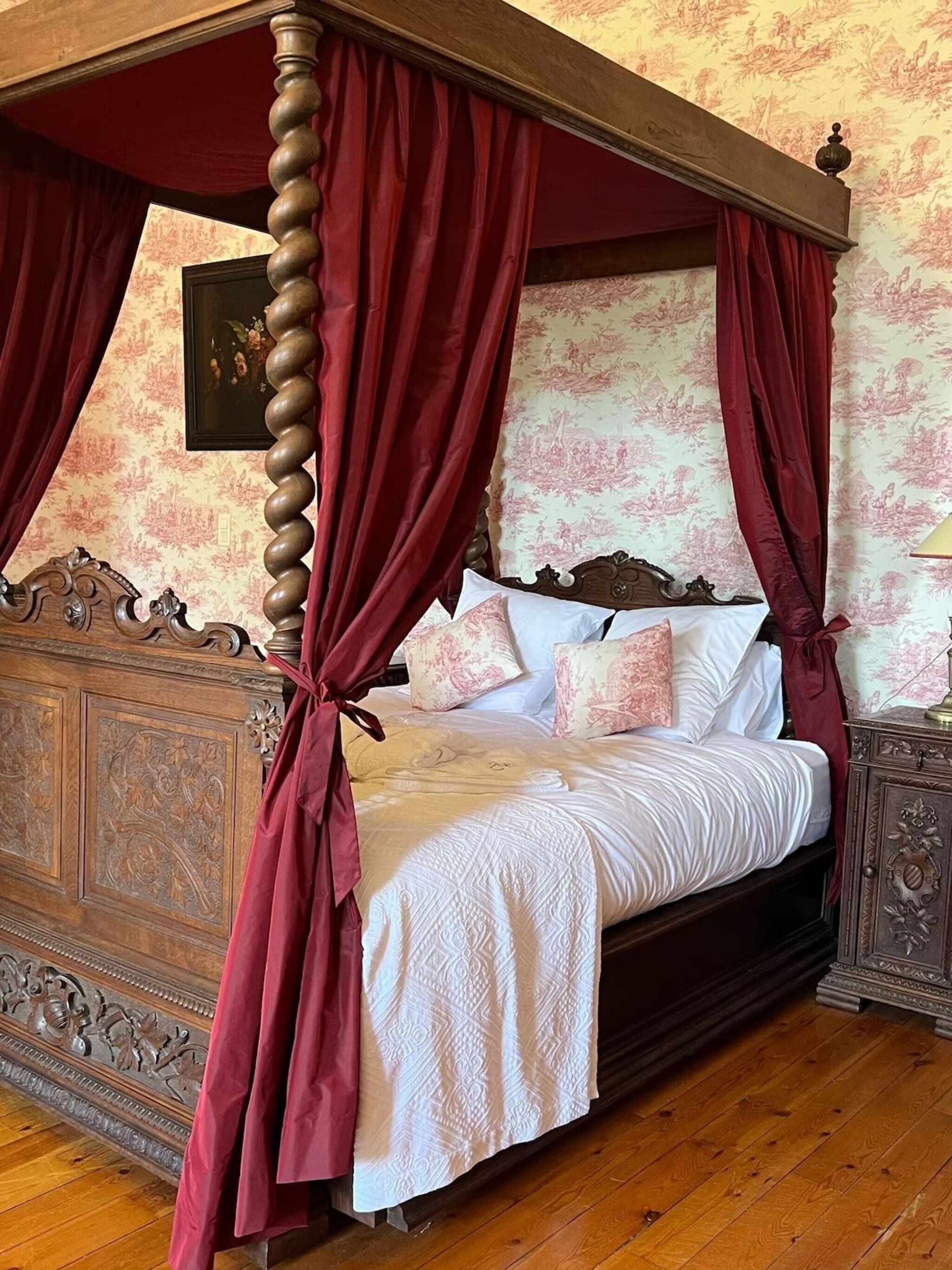 A four-poster canopy bed with red curtains, a wooden headboard, and floral-patterned wallpaper.