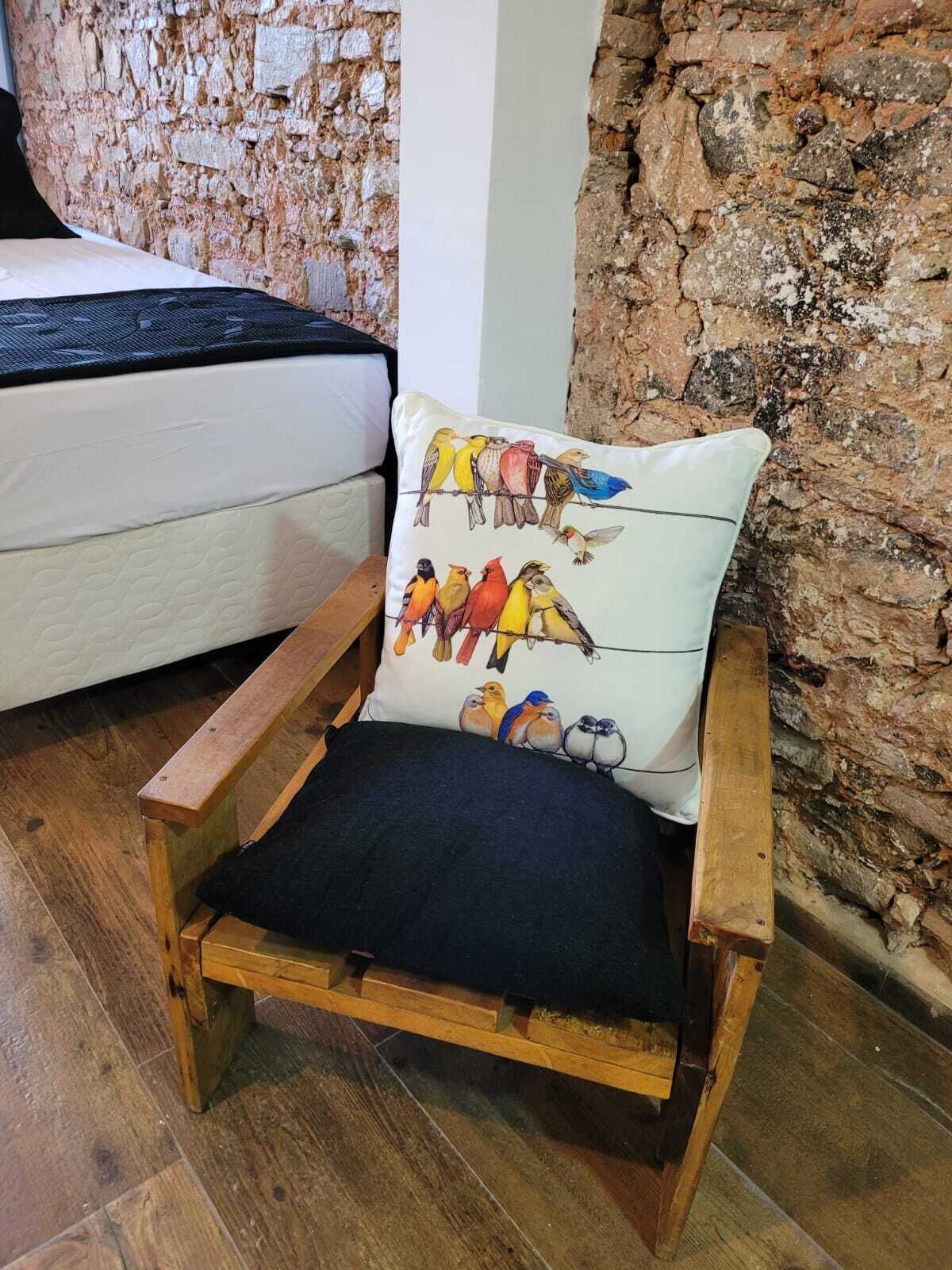 A wooden chair with a cushion featuring colorful birds, positioned in a room with a stone wall and a white sofa.