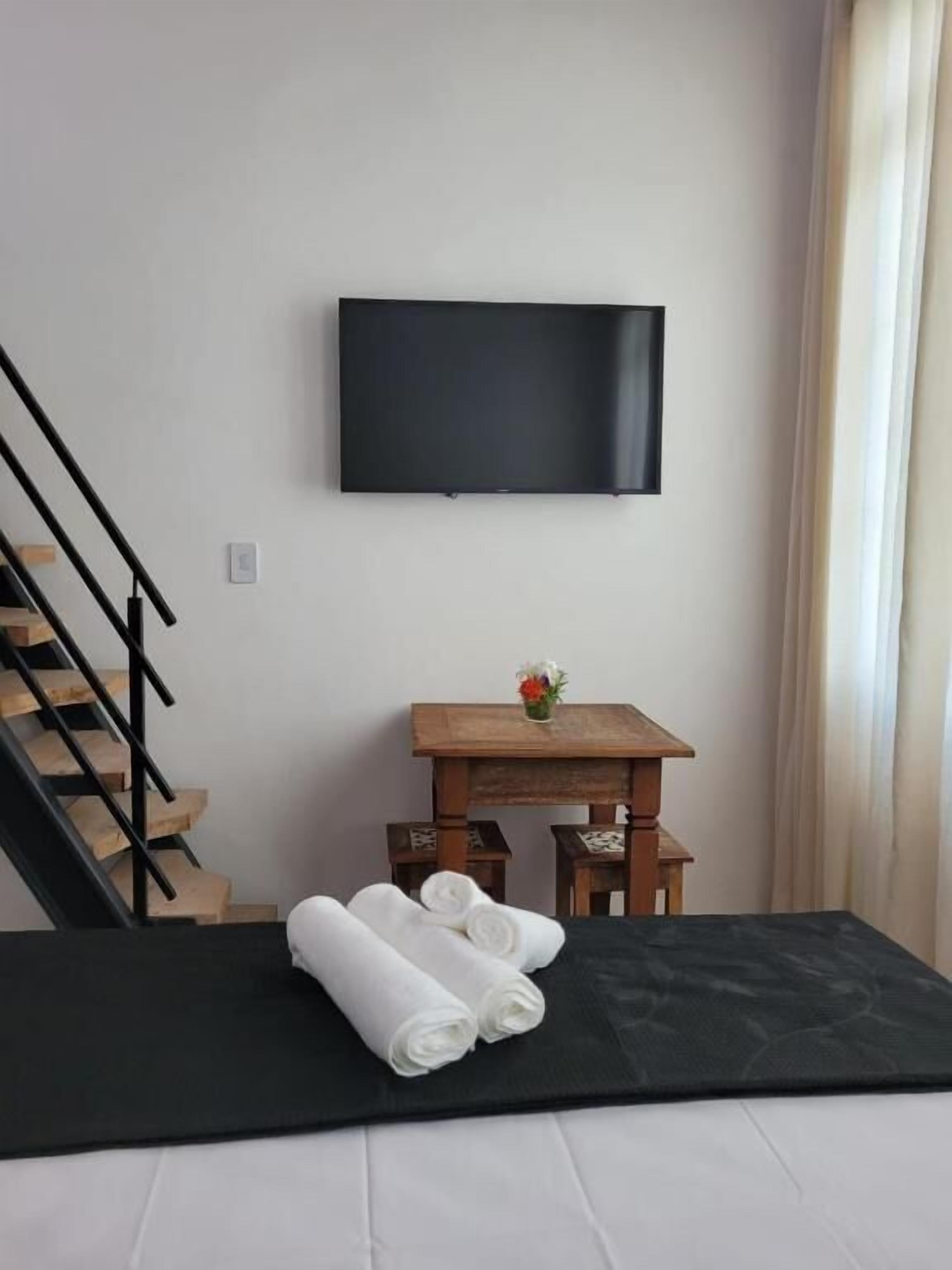 Ein Zimmer mit einem Bett, einem an der Wand befestigten Fernseher, einem kleinen Holztisch mit Blumenvase und einer Treppe.