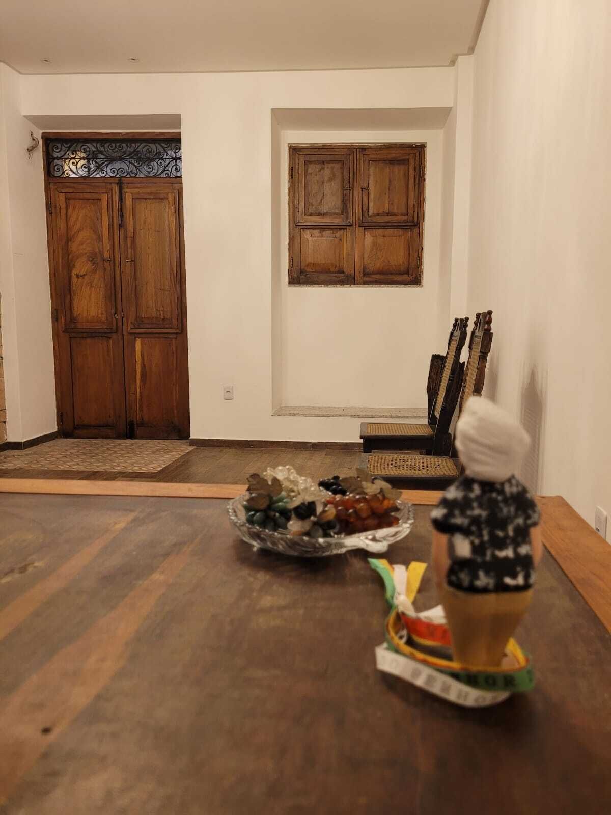 Um quarto com porta de madeira, uma janela com persianas de madeira, uma mesa de jantar com um recipiente de petiscos e uma guitarra encostada na parede.