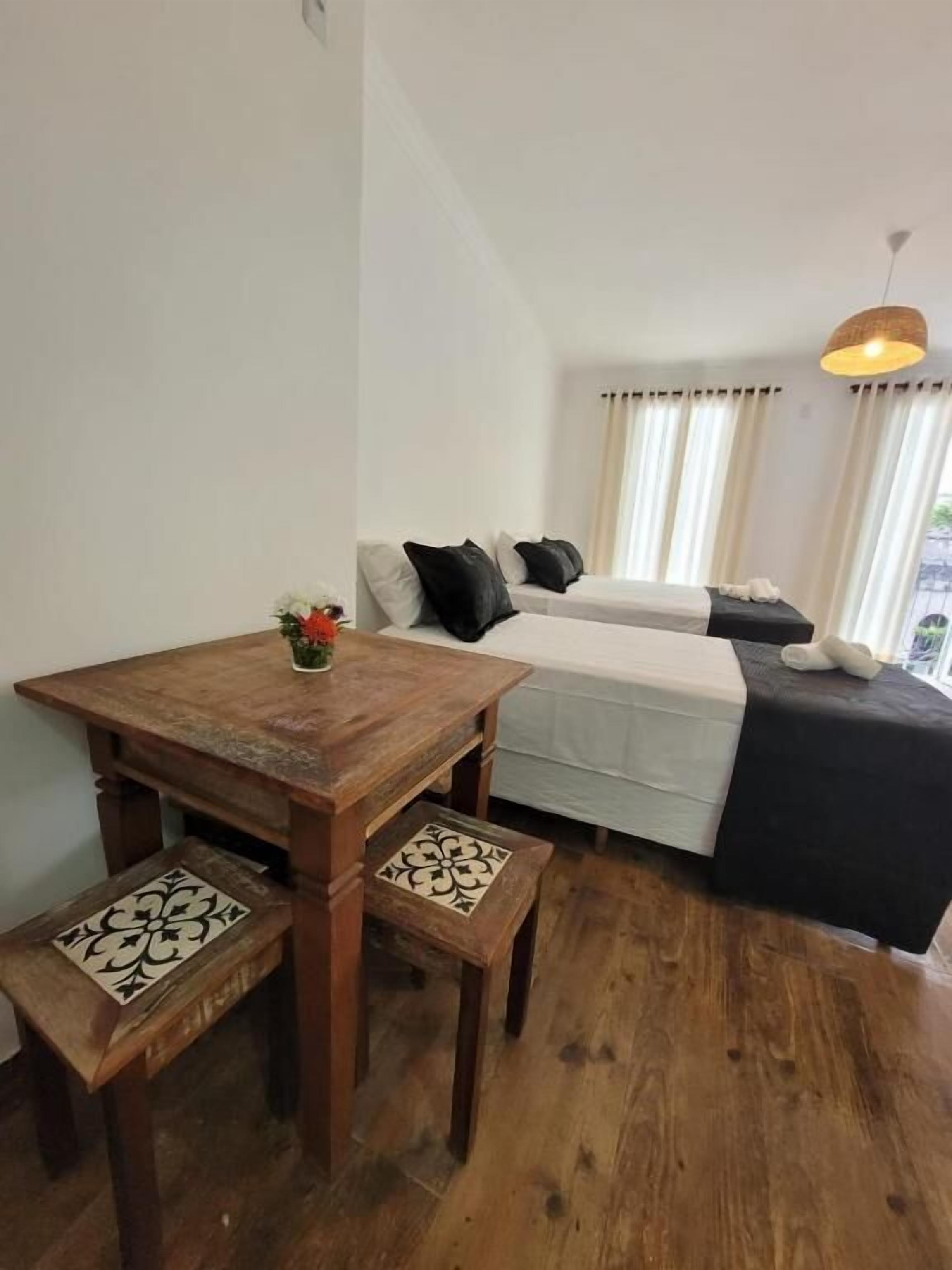 A small, cozy room with a wooden table and benches, a bed with a white and black bedspread, and a hanging light fixture.