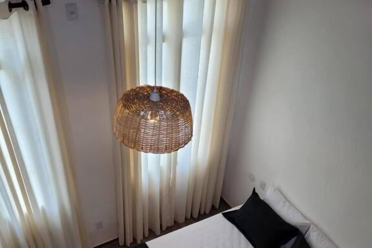 A neatly made bed with a black and white bedspread, white pillows, and a rolled towel on the side. There is a wicker lamp hanging above the bed, and white curtains are drawn to the sides.