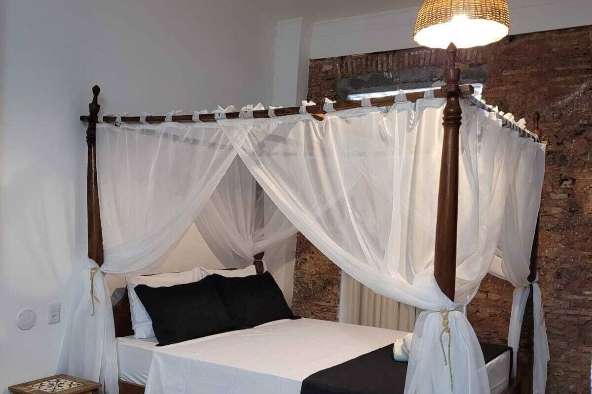 A four-poster bed with white bedding and a canopy, a small wooden nightstand, and a brick wall in the background.