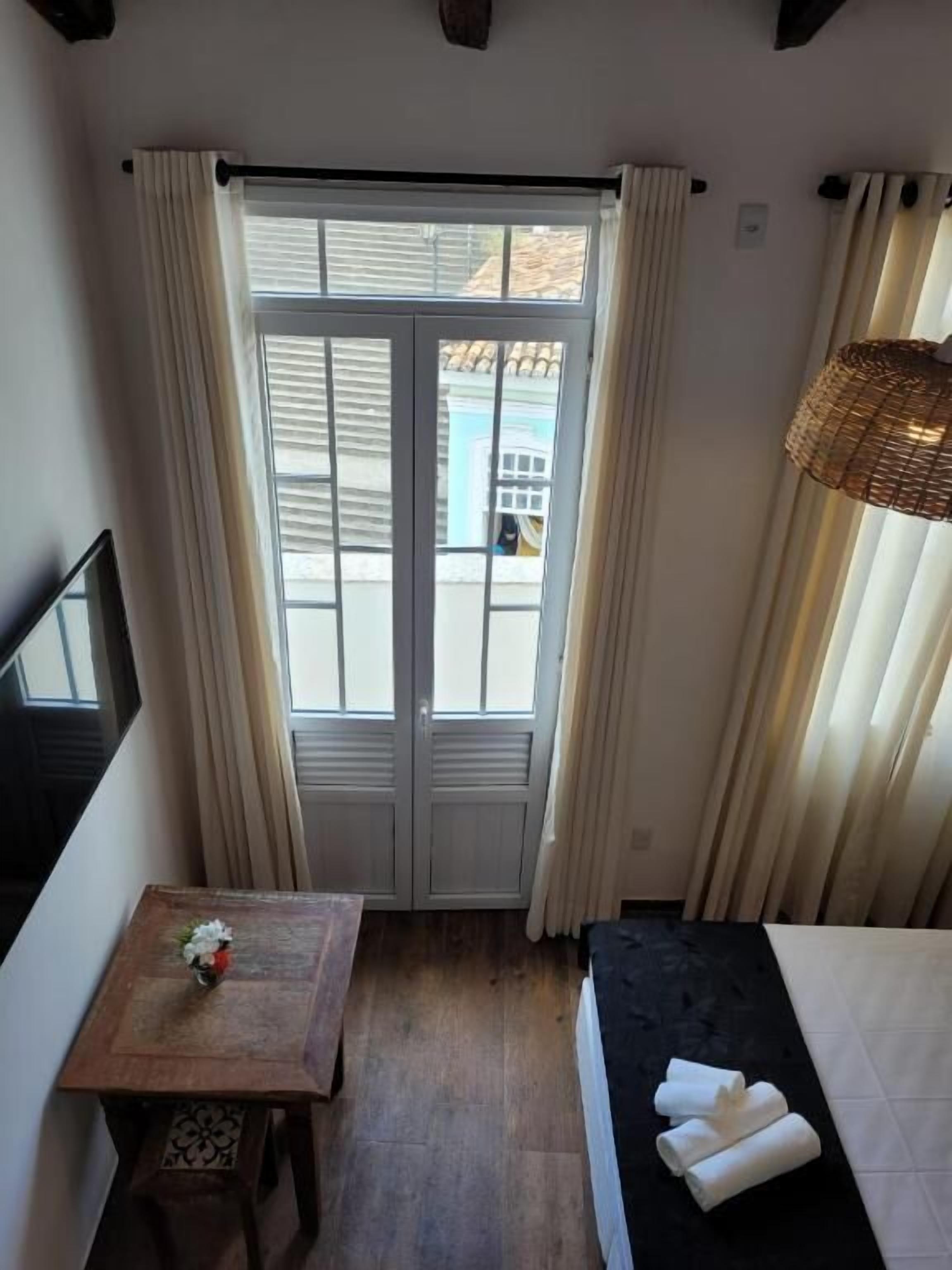 Una habitación con una cama, una mesita con una flor, una puerta con cortinas y vistas al exterior.