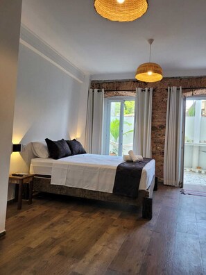 A bedroom with a large bed, a nightstand, a hanging light fixture, and a view of the outside through a glass door. - Pelourinho Boutique Hotel - OH HOTÉIS (Salvador)