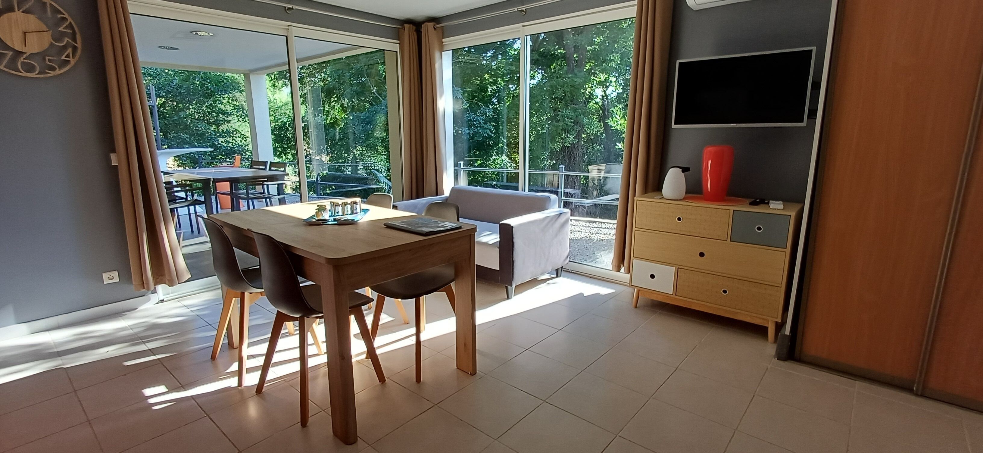 Une pièce moderne comprenant une table à manger, des chaises, un canapé, un téléviseur et une grande fenêtre donnant sur des arbres.