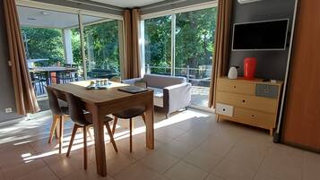 Une pièce moderne comprenant une table à manger, des chaises, un canapé, un téléviseur et une grande fenêtre donnant sur des arbres.