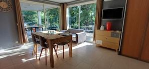 Une pièce moderne comprenant une table à manger, des chaises, un canapé, un téléviseur et une grande fenêtre donnant sur des arbres.