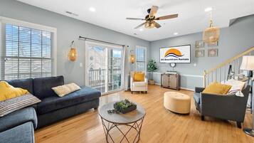 A spacious living room with a grey sofa, a round coffee table, and a grey armchair. There is a ceiling fan, a chandelier, and a large window with blinds.