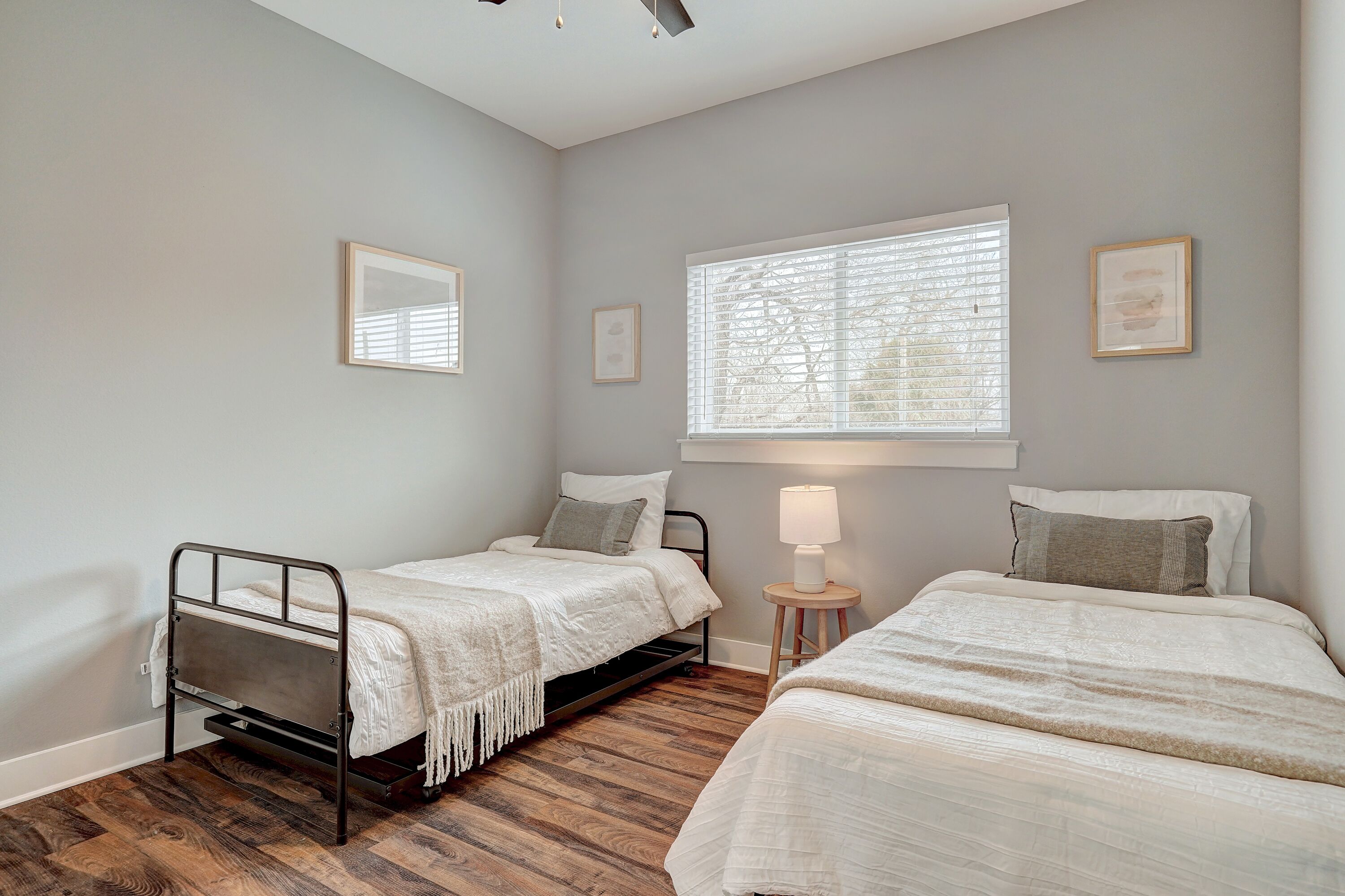A bedroom with two beds, a window with blinds, a bedside lamp, and framed pictures on the wall.