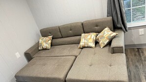 Un sofá modular con tela gris, dos cojines estampados y una ventana con cortinas.