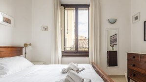 Una cama bien tendida con sábanas blancas y toallas enrolladas, un cabecero de madera, una mesita de noche con lámpara, un aparador y una ventana con cortinas.