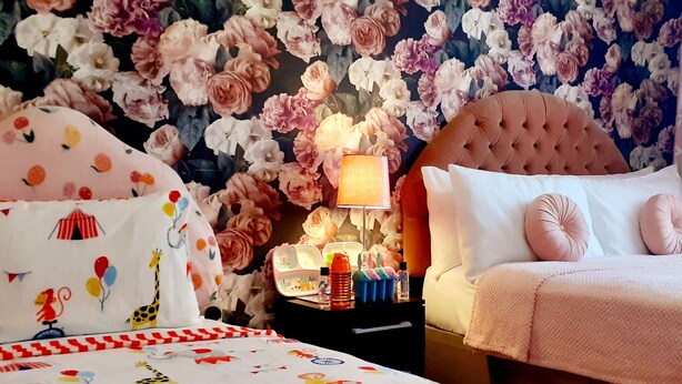 Ein Schlafzimmer mit floraler Tapete, einem gepolsterten Kopfteil, einem Nachttisch mit Lampe und einem Bett mit einer rosa Decke.