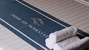 A bed with a blue cover featuring the text "PHAM GIA HOMESTAY" and a logo. There are two rolled white towels on the bed.