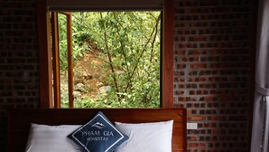 A bed with white linens and a pillow with "PHAM GIA HOMESTAY" text, a brick wall, and a window with a view of greenery.