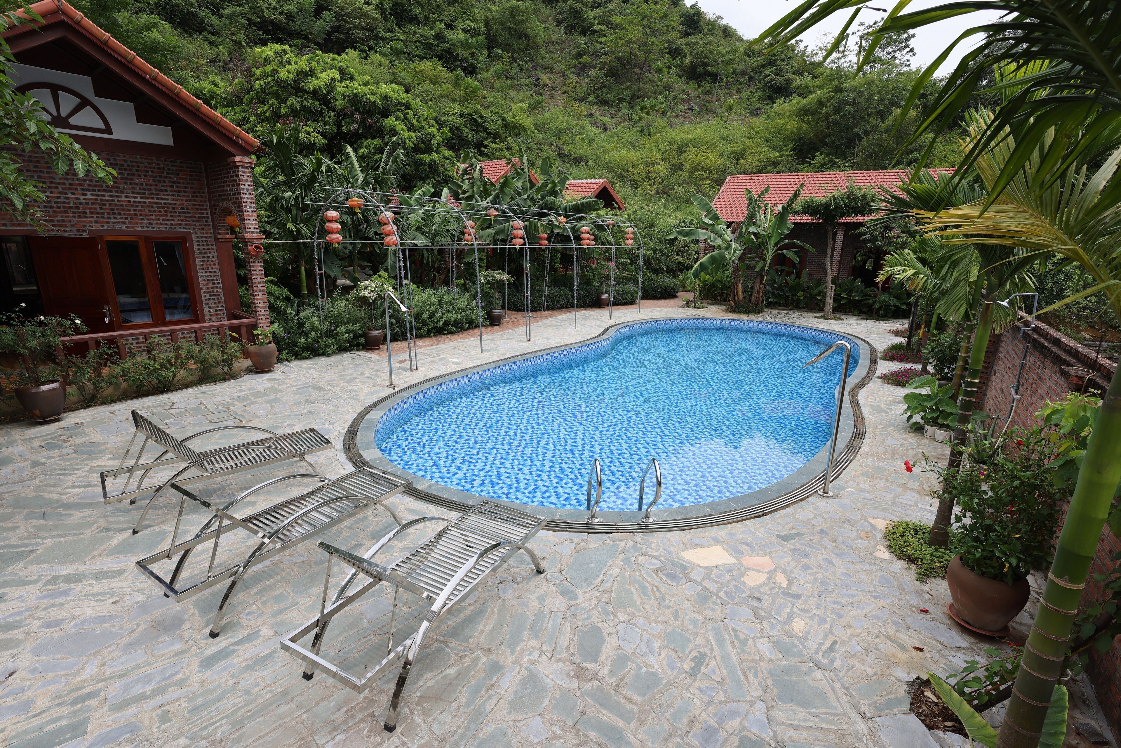 A swimming pool with stone tiles, metal loungers, and a brick building with a red roof.