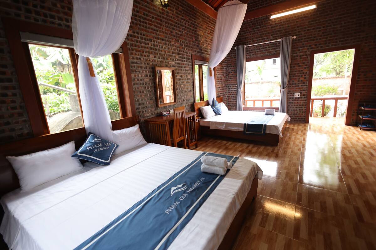 A hotel room with a large bed, a smaller bed, and a seating area. There are windows with curtains, a brick wall, and a wooden floor.