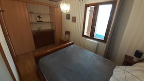 A compact room with a bed, a small desk, a chair, a shelf with decorative items, a window with curtains, and a hanging light fixture. - Country house close to town: 1 star (SARLAT)