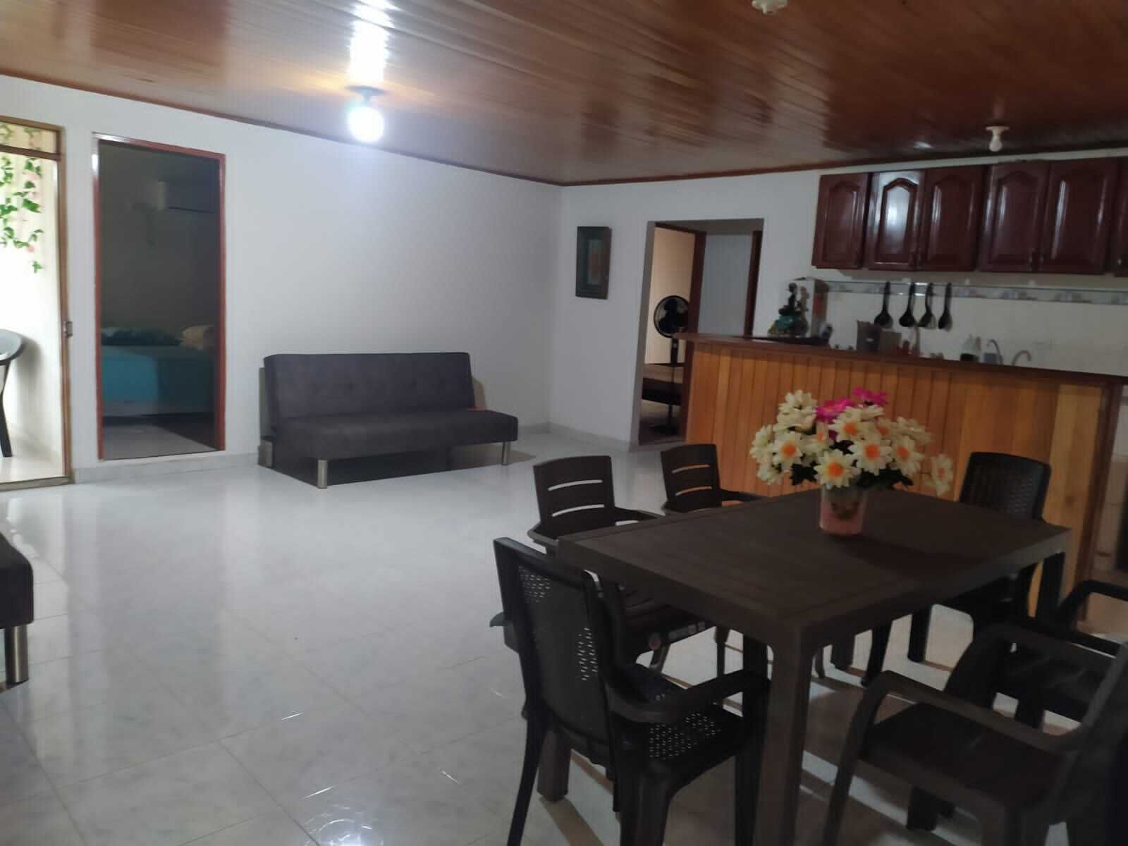 A spacious living area with a dark sofa, a dining table with chairs, and a kitchen area with wooden cabinets.