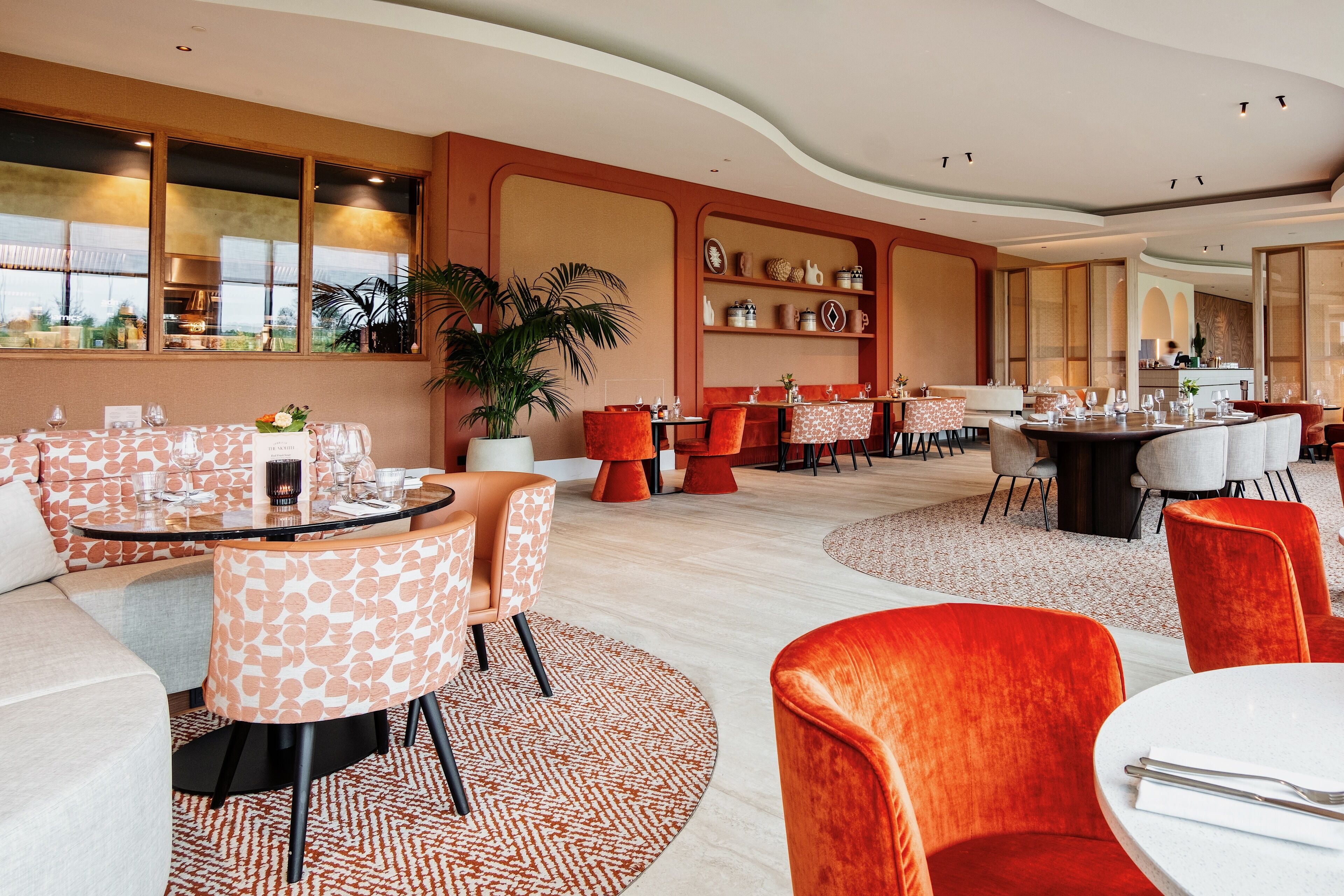 Un restaurante moderno con mesas redondas, sillas anaranjadas y blancas, y una gran planta en maceta.