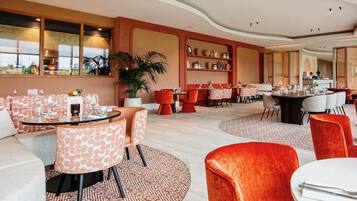 Un restaurante moderno con mesas redondas, sillas anaranjadas y blancas, y una gran planta en maceta.